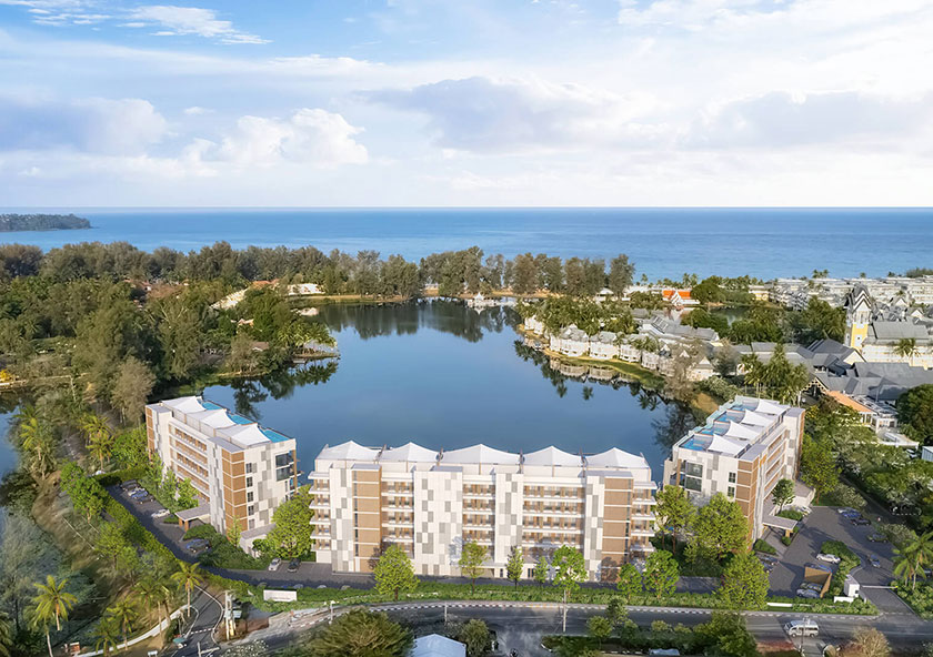 Angsana Oceanview Residences Phuket | An aerial view of modern buildings beside a calm lake with a backdrop of the ocean and a clear blue sky.