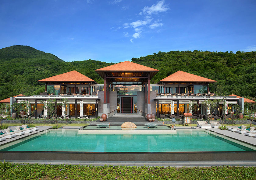 Banyan Tree Residences Lăng Cô | An elegant resort with a large swimming pool in the foreground, set against a backdrop of lush green hills under a clear blue sky.