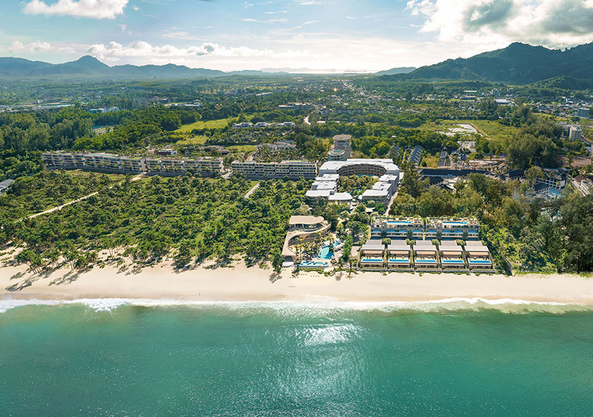 Banyan Tree Residences Beach Residences | Aerial view of a coastal resort with multiple buildings nestled among greenery beside a sandy beach, with a backdrop of mountains.