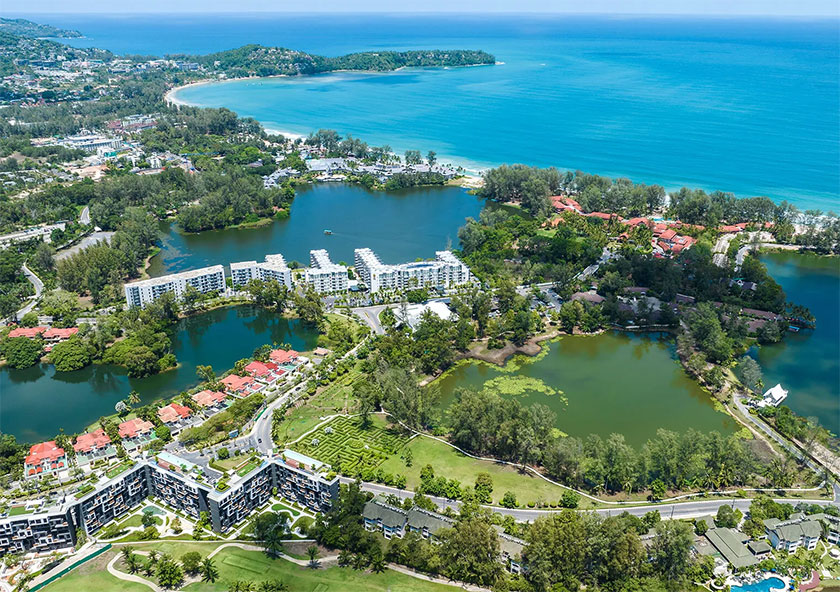 Cassia Phuket | An aerial view captures a coastal area with resorts, a lake, abundant greenery, and a beach edging a clear blue sea.