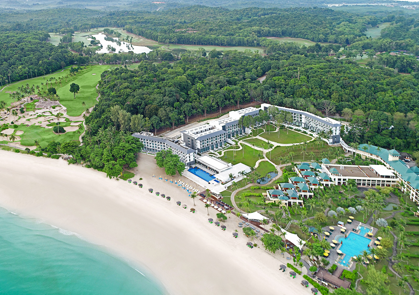 Cassia Residences Bintan | Aerial view of a beachfront resort beside a golf course surrounded by dense foliage.