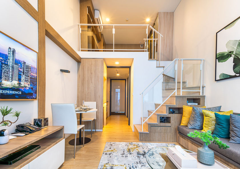 Cassia Residences Rama 9 | Modern, well-lit interior of a compact apartment with a wooden staircase leading to an upper level, a couch with colorful cushions.
