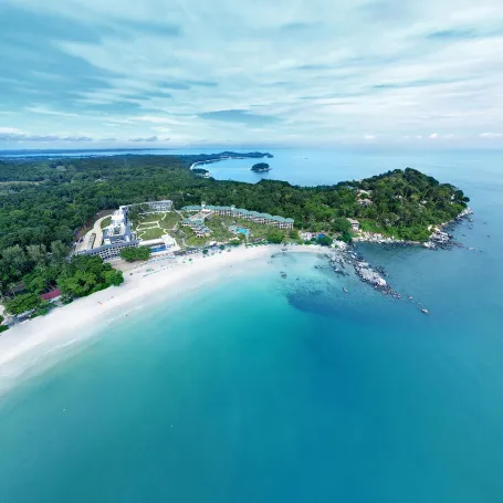 Cassia Residences Bintan | Aerial view of a coastal resort with white buildings and pools next to a turquoise sea, bordered by a lush green forest.