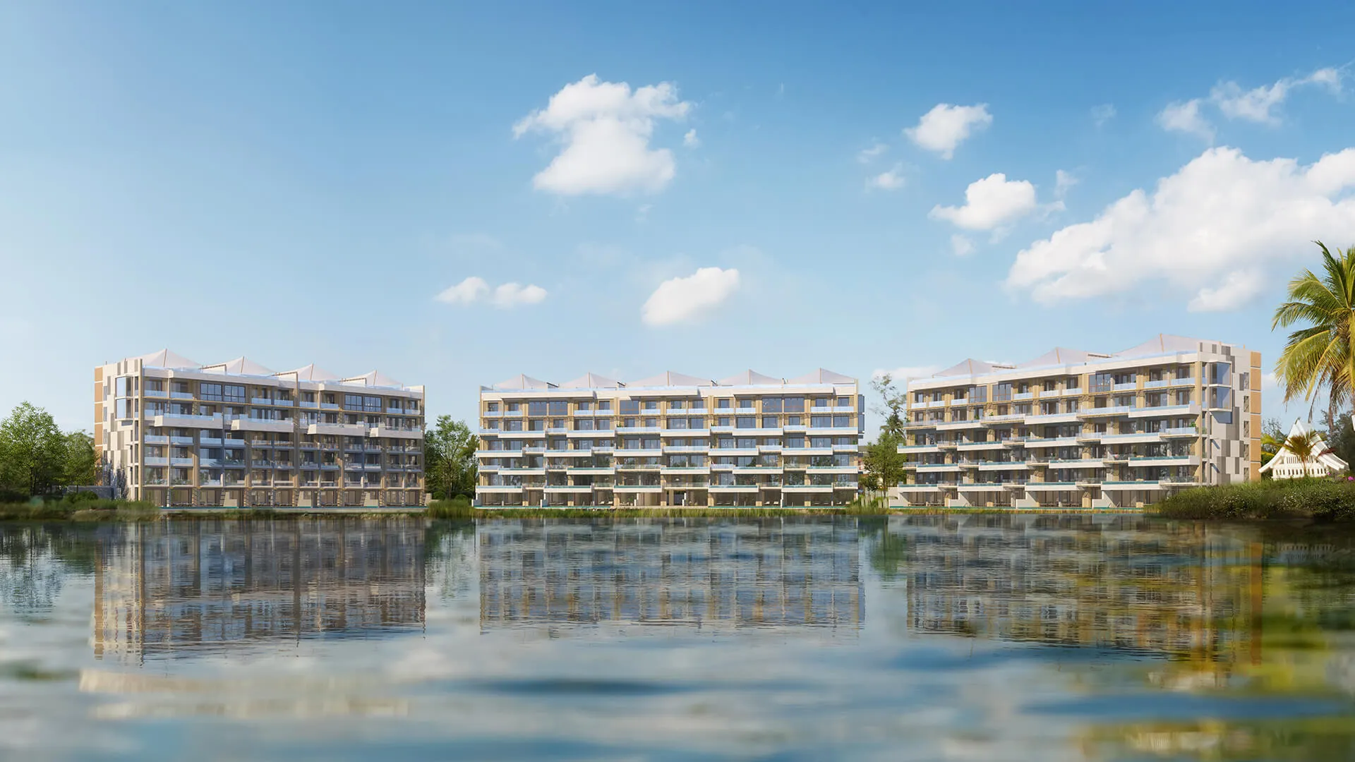 Symmetrical modern apartment buildings with large balconies stand beside a calm body of water, under a clear blue sky with clouds.