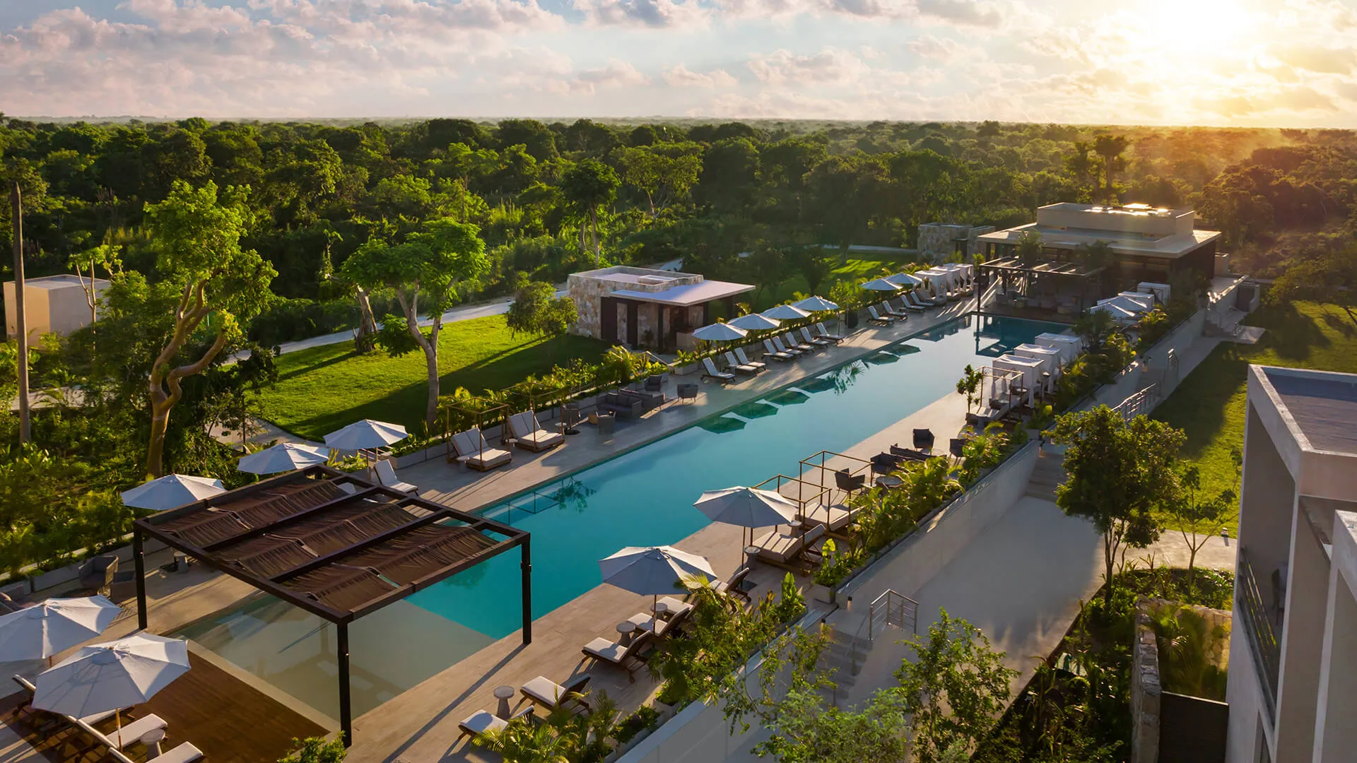 Angsana Hacienda Xcanatun - A luxurious pool stretches alongside loungers and cabanas, surrounded by verdant foliage and buildings, under a bright, setting sun.