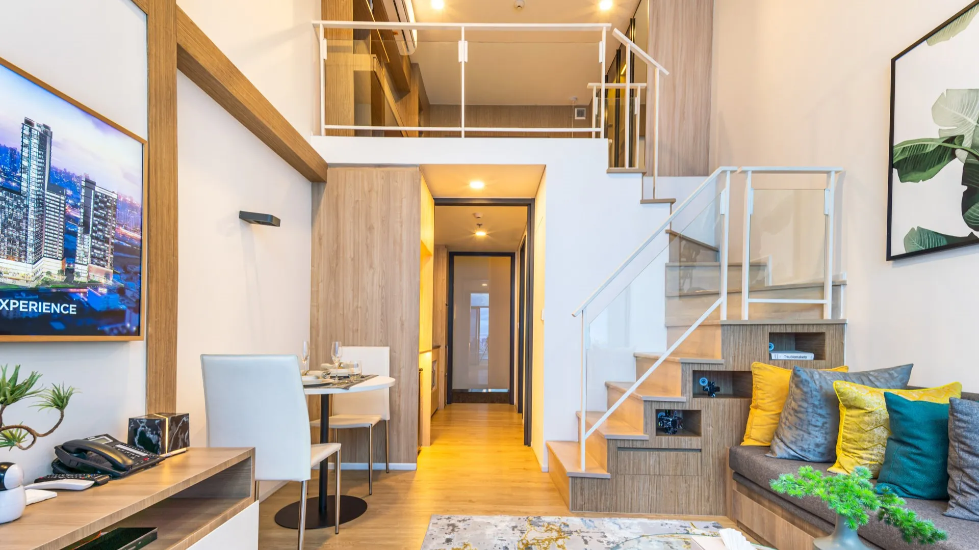 Cassia Residences Rama 9 - Modern living room with a sofa, dining area, and a TV, leading to a wooden staircase and an upper level.